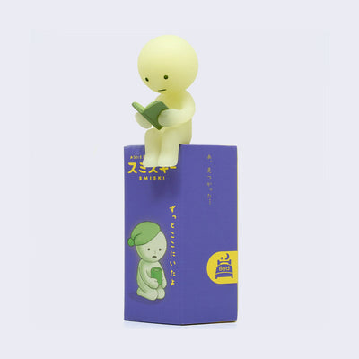 Small light green simple character with a round head, reading a book. It sits atop of its purple blind box packaging.