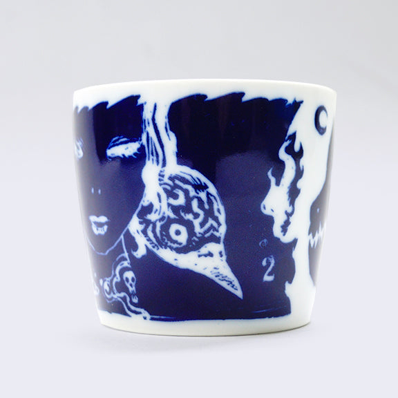 White ceramic cup with a wrap around dark blue illustration. Current side features a close up of a woman's face with a bird's head peeping over her shoulder and the number 2.