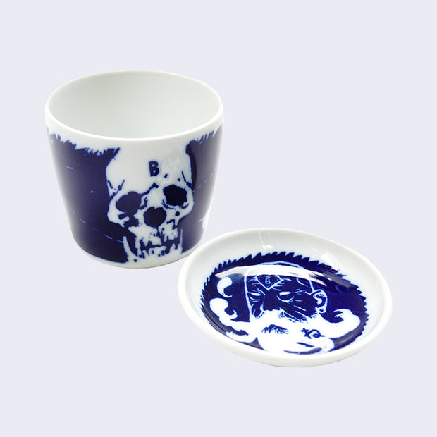 White ceramic cup and saucer set, with dark blue illustration on both. Cup features a wrap around illustration, with the current side featuring a skull. The saucer plate in front features an old bearded man.