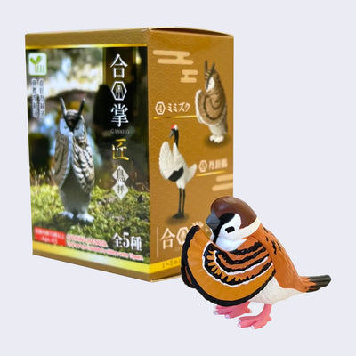 Small plastic sparrow figure, with its eyes closed, head slightly bowed and wings pressed together in a prayer pose. Behind it is its blind box packaging, brown with photos of other bowing birds. 