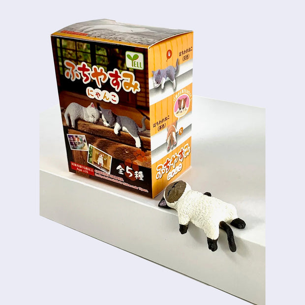 Small plastic figure of a Siamese cat, laying on its back with its limbs draped over the edge of a ledge. It stands next to a box with photos of the other design options and Kanji text.