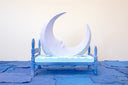 Carved wooden sculpture of a large purple crescent moon with a small face, resting on a flat blue bed on a purplish blue wooden bedframe. 