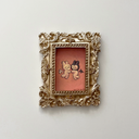 Small painting of 2 bunny characters with facial features made of lines, like emotes, both with a sad face. They float and face one another, holding hands. One is dressed as an angel, the other a demon. Piece is in a very ornate golden frame.