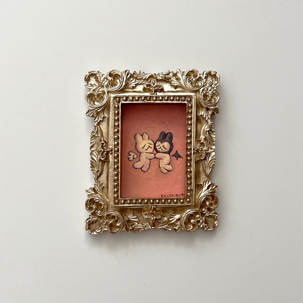 Small painting of 2 bunny characters with facial features made of lines, like emotes, both with a sad face. They float and face one another, holding hands. One is dressed as an angel, the other a demon. Piece is in a very ornate golden frame.