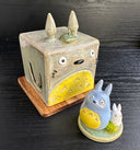 Set of ceramic sculptures, one cube shaped and modeled to look like Totoro. Nearby are a small blue and white chibi Totoro, standing on a small circle platform with a grass tuft.