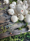 Mixed media sculpture of many small cute white blob creatures emerging from wooden floor boards with a large hole in the middle and greenery all around. Piece is within a black shadowbox frame without glass.