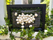 Mixed media sculpture of many small cute white blob creatures emerging from wooden floor boards with a large hole in the middle and greenery all around. Piece is within a black shadowbox frame without glass.