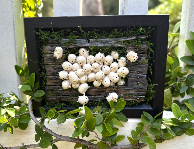 Mixed media sculpture of many small cute white blob creatures emerging from wooden floor boards with a large hole in the middle and greenery all around. Piece is within a black shadowbox frame without glass.