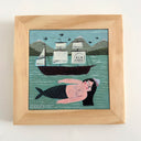 A framed folk art painting depicting a mermaid woman with black hair floating in teal water under to a dark boat, with mountains in the background, housed in a light wood frame.