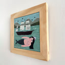A framed folk art painting depicting a mermaid woman with black hair floating in teal water under to a dark boat, with mountains in the background, housed in a light wood frame.