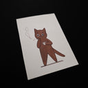 Artwork featuring a hand-drawn brown cartoon cat holding a small white cup and smoking a cigarette, photographed at an angle on a dark black background.