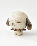 A small speckled ceramic figurine of a dog-like character with closed eyes, brown floppy ears, and a pink spiked collar, centered on a plain white background.