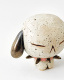 A small speckled ceramic figurine of a dog-like character with closed eyes, brown floppy ears, and a pink spiked collar, centered on a plain white background.