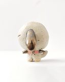 A small speckled ceramic figurine of a dog-like character with closed eyes, brown floppy ears, and a pink spiked collar, centered on a plain white background.