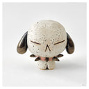A handmade ceramic figurine of a round, speckled white dog with dark brown ears and a small pink collar, photographed on a neutral light gray background.