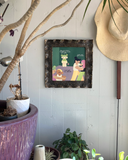 A square ceramic frame with textured, speckled dark brown and black glaze. It frames a colorful illustration of characters dancing and standing on a green background, with text accompanying it. Hung in an indoor garden setting.