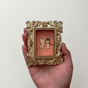 Small painting of 2 bunny characters with facial features made of lines, like emotes, both with a sad face. They float and face one another, holding hands. One is dressed as an angel, the other a demon. Piece is in a very ornate golden frame.