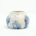 Cream colored vase with blue flowers as a repeating design, they have grid patterning and their coloring bleeds down the side of the cup. Gold specks decorate the cup.