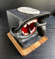 Underside of a black ceramic cube, with a large open mouth with 2 hands going towards it, as if throwing food into it. Top of the cube has a "No Face" mask from Spirited Away.