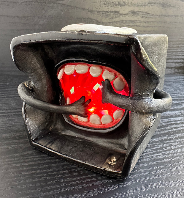 Underside of a black ceramic cube, with a large open mouth with 2 hands going towards it, as if throwing food into it. Top of the cube has a "No Face" mask from Spirited Away.