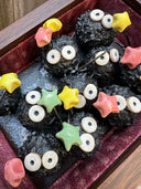 Diorama style sculptural work of many black soot sprites holding colorful stars. They're within a wooden box with red fabric lining it.