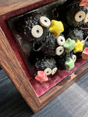 Diorama style sculptural work of many black soot sprites holding colorful stars. They're within a wooden box with red fabric lining it.