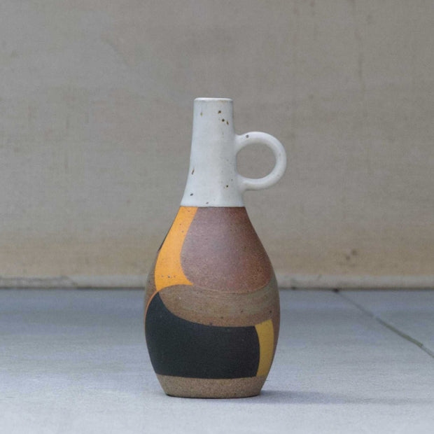 Ceramic pot with a narrow, tall opening and a small round handle. Top half is white and speckled, bottom half consists of geometric shapes painted brown, orange and black.