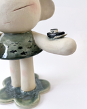 A handmade ceramic figurine of a stylized girl with a crown, short hair like ocean waves, and her hand extended out, with a tiny sailboat on her arm. Her dress has a fish pattern.