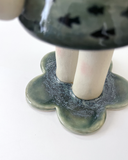 A handmade ceramic figurine of a stylized girl with a crown, short hair like ocean waves, and her hand extended out, with a tiny sailboat on her arm. Her dress has a fish pattern.