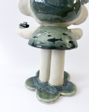A handmade ceramic figurine of a stylized girl with a crown, short hair like ocean waves, and her hand extended out, with a tiny sailboat on her arm. Her dress has a fish pattern.