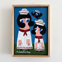 A framed folk art painting of two stylized figures in white sailor dresses with red bows, a black bird flying over them with a matching sailor hat, and the name 'addeline' written at the bottom, displayed on a light wall.