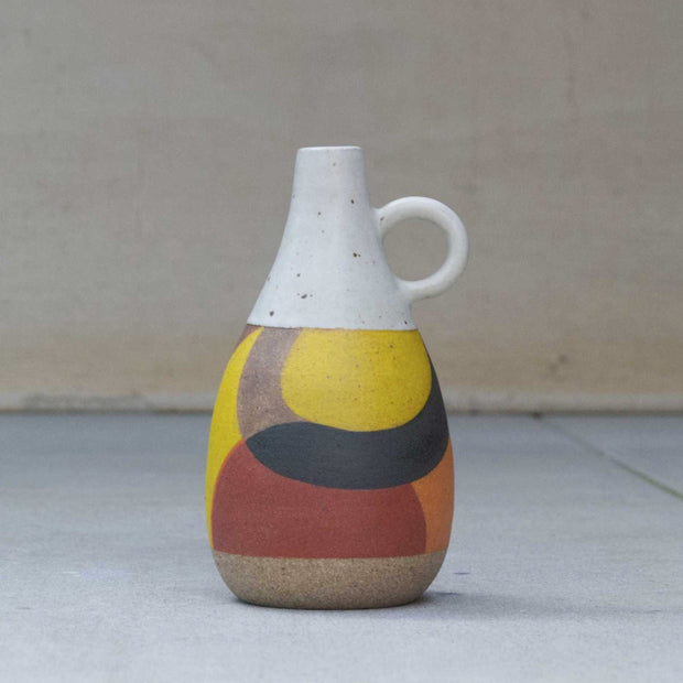 Ceramic pot with a narrow, tall opening and a small round handle. Top half is white and speckled, bottom half consists of geometric shapes painted yellow, brown, red and orange.