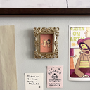 Small painting of 2 bunny characters with facial features made of lines, like emotes, both with a sad face. They float and face one another, holding hands. One is dressed as an angel, the other a demon. Piece is in a very ornate golden frame. Hung on a wall with notes around it.