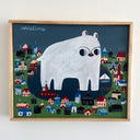 A framed hand-painted illustration of a giant white polar bear, standing over a colorful coastal town. The artwork is housed in a light natural wood frame and hangs on a plain white wall.