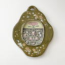 An organic-shaped ceramic frame with a textured olive green glaze, featuring a central oval inset illustration with a stylized pink flower and a detailed root system underneath with girls dancing around.