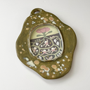 An organic-shaped ceramic frame with a textured olive green glaze, featuring a central oval inset illustration with a stylized pink flower and a detailed root system underneath with girls dancing around.