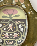 An organic-shaped ceramic frame with a textured olive green glaze, featuring a central oval inset illustration with a stylized pink flower and a detailed root system underneath with girls dancing around.