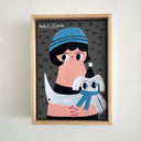 A framed hand-painted illustration on a dark gray background, showing a stylized person in a blue beanie holding a white dog with a blue scarf. The artwork is housed in a light natural wood frame and hangs on a white wall.
