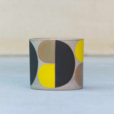 Short ceramic cup, without any curvature. The natural light tan clay color shows through with large painted geometric shapes over it in brown, black and yellow.
