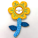 A hand-painted wooden flower decoration with yellow petals, a smiling white center, blue and brown details, and a blue stem with white dots and leaf shapes, set against a plain white background.