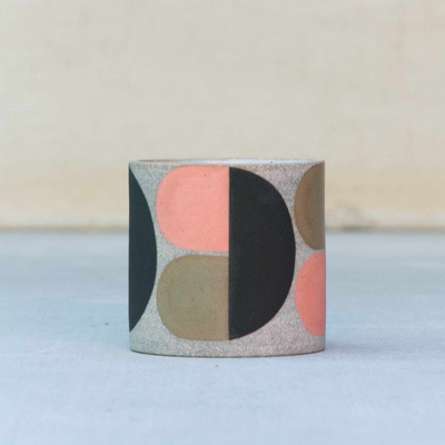 Short ceramic cup, without any curvature. The natural light tan clay color shows through with large painted geometric shapes over it in pink, brown and black.