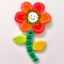 Flat, colorful clay sculpture of a red flower with a green patterned stem and a white smiling face in the center.