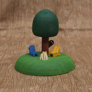 Sculptural piece of a round tree on a mound of grass. In front are 2 wooden chairs and an unlit bonfire. Behind the tree is a shadowy figure, peeking out.