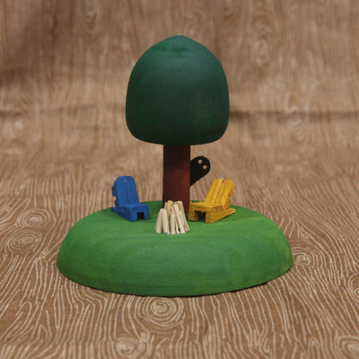 Sculptural piece of a round tree on a mound of grass. In front are 2 wooden chairs and an unlit bonfire. Behind the tree is a shadowy figure, peeking out.