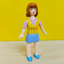 Clay figure of a woman with short brown hair and bangs, standing with her arms slightly out at her sides and wearing an office outfit - a blue skirt with a pink button up short sleeve shirt and yellow plaid vest.