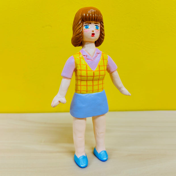 Clay figure of a woman with short brown hair and bangs, standing with her arms slightly out at her sides and wearing an office outfit - a blue skirt with a pink button up short sleeve shirt and yellow plaid vest.