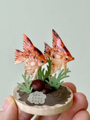 Two small fish figurines on a circular base with greenery, held by a hand against a light gray background.