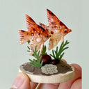 Decorative fish figurine on a small base with greenery, held by a hand against a light background.