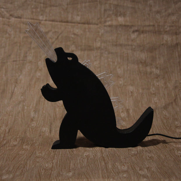 Die cut wooden silhouette of Godzilla, made into a lamp. A beam of light comes out of its mouth and it has lit up spikes along its back. Lamp is shown off.