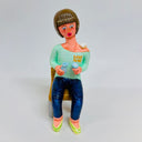 Clay sculpture of a woman with short brown hair and bangs, sitting on a chair pouring herself a cup of sake. Resting on her shoulder is a small square object, possibly a piece of food or a folded towel.